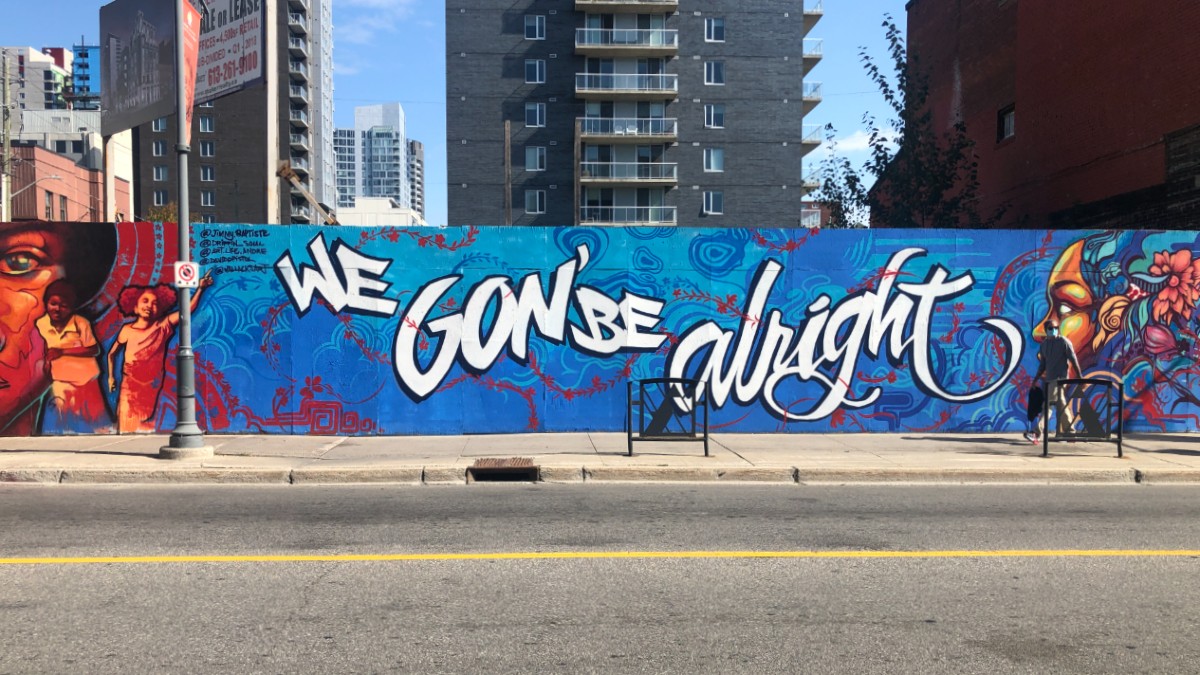 A long mural is shown from across the street. The words "We Gon' Be Alright" in white stretch across a blue background. On either side of the words are women painted in golden tones.