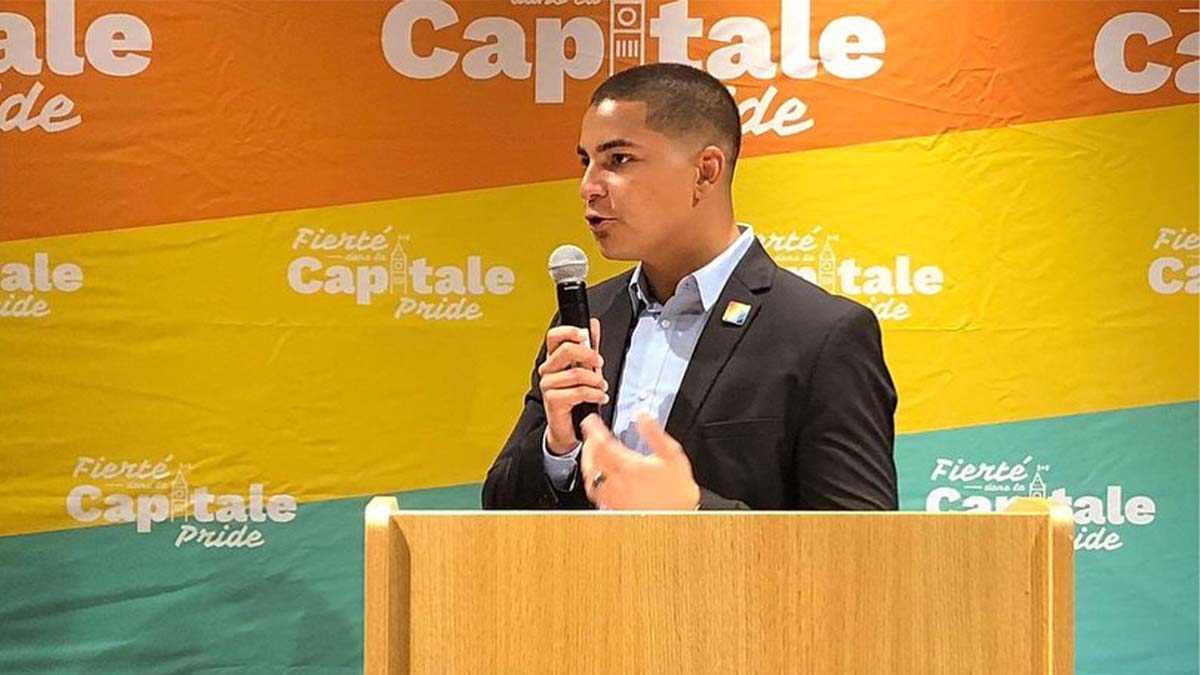 A photo of Davy Sabourin, Capital Pride’s chairperson on the Board of Directors standing at a podium and holding a microphone behind a Capital Pride banner.