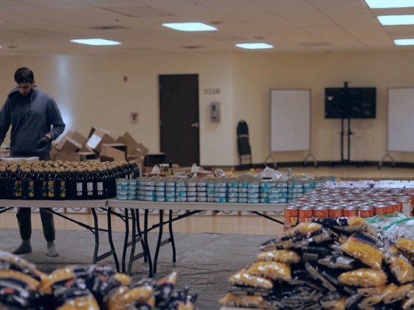 Momin Janjua stands at a table and is packaging boxes, with food supplies in the back and foreground. 