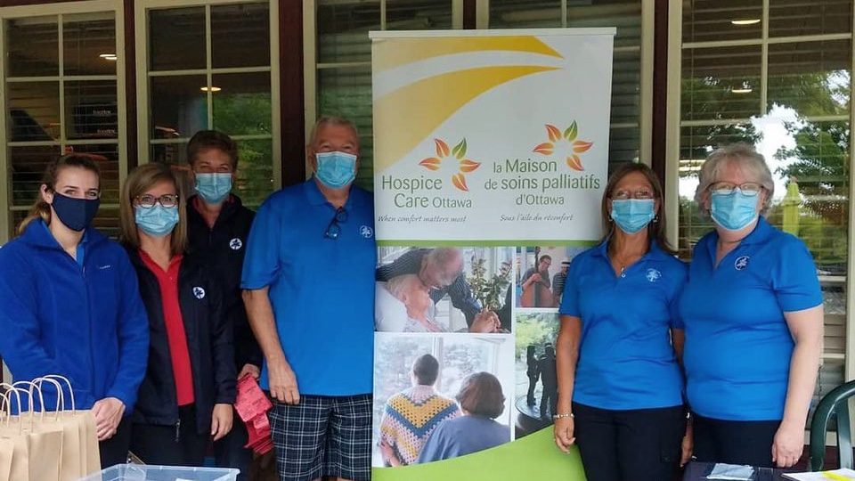 Six people stand in front of a Hospice Care Ottawa banner. They are wearing masks and blue shirts.