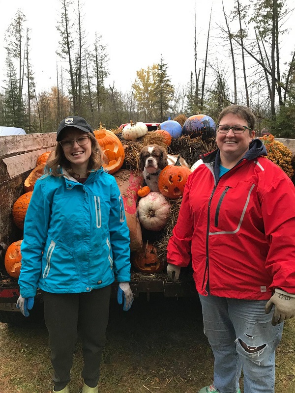 Two women standing next to each other in front of a truck filled with pumpkins.