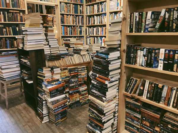 Walls lined with floor to ceiling bookshelves full of books, and other tables and shelves in the centre, as well as piles of books stacked high.