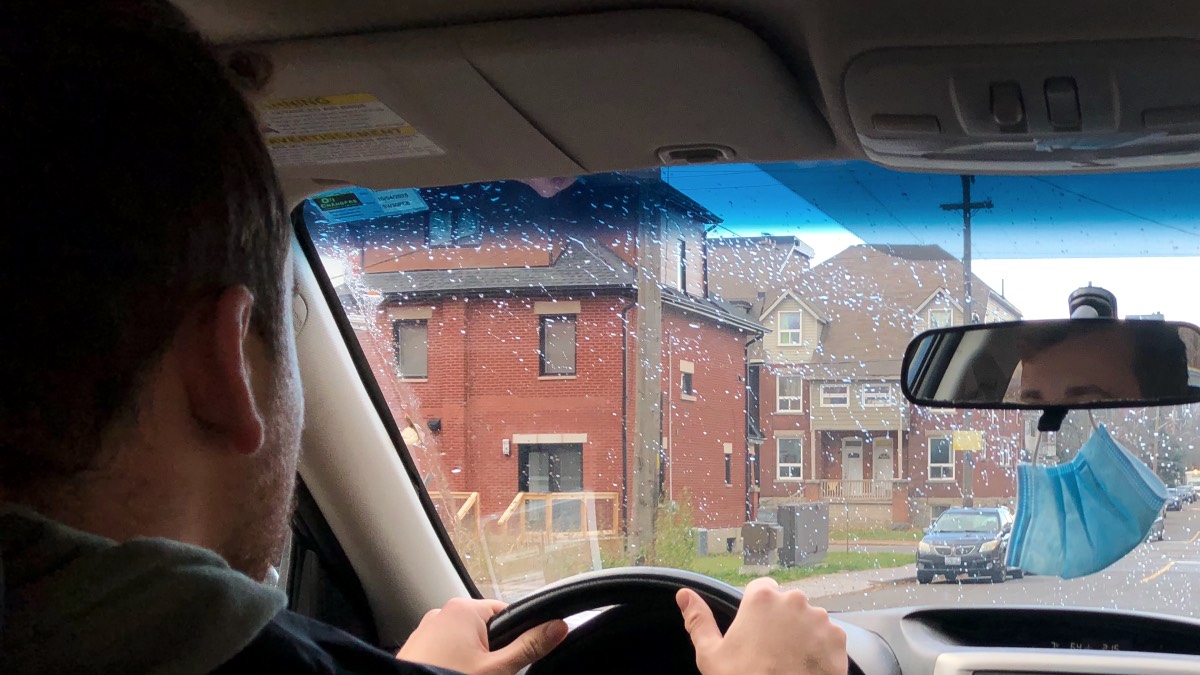 A man driving a car as seen from backseat. A blue mask is hanging from the rear-view mirror. A residential area is displayed through the windshield.