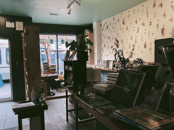 Front nook of Black Squirrel Books with tables at different heights as well as bookshelves, all covered with books and a wall of hanging dried flowers. Hand sanitizer also pictured near the entrance.