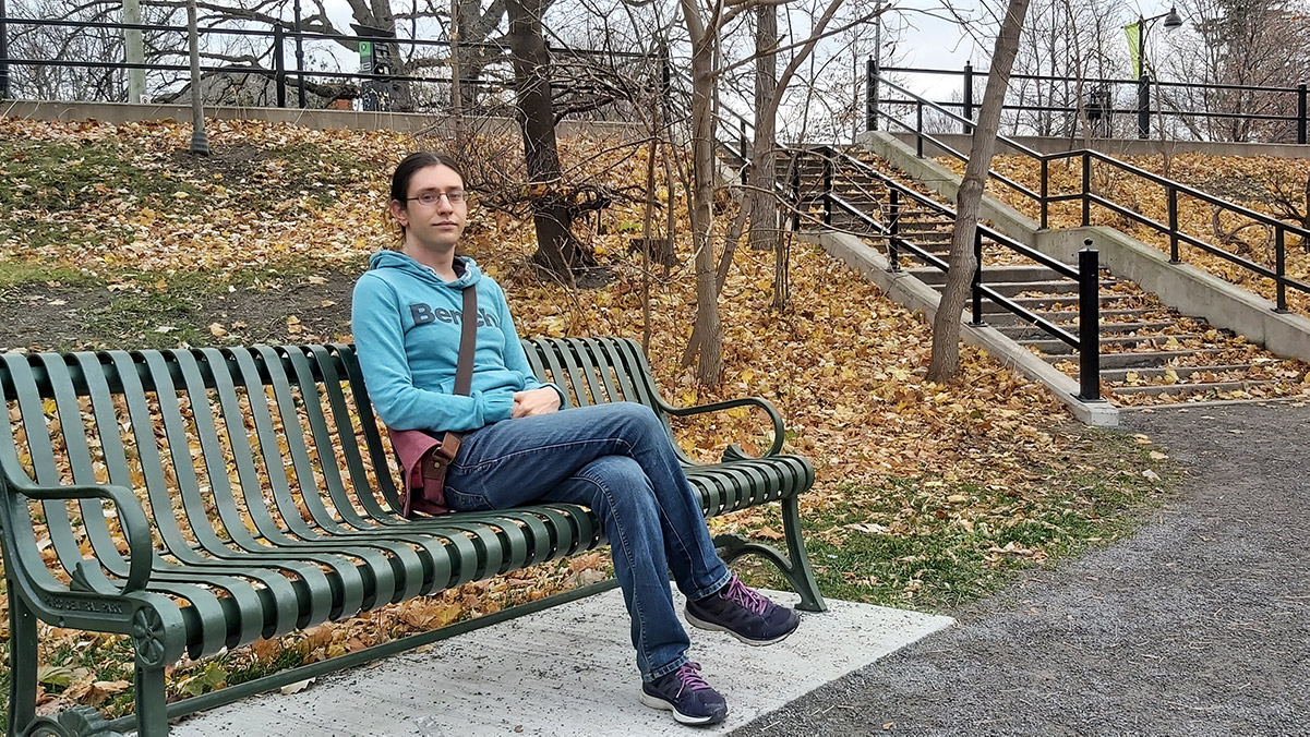 Lyra Evans is sitting on a green park bench. Behind her are fall leaves and the stairs up to Bank St. in Patterson's Creek Park.