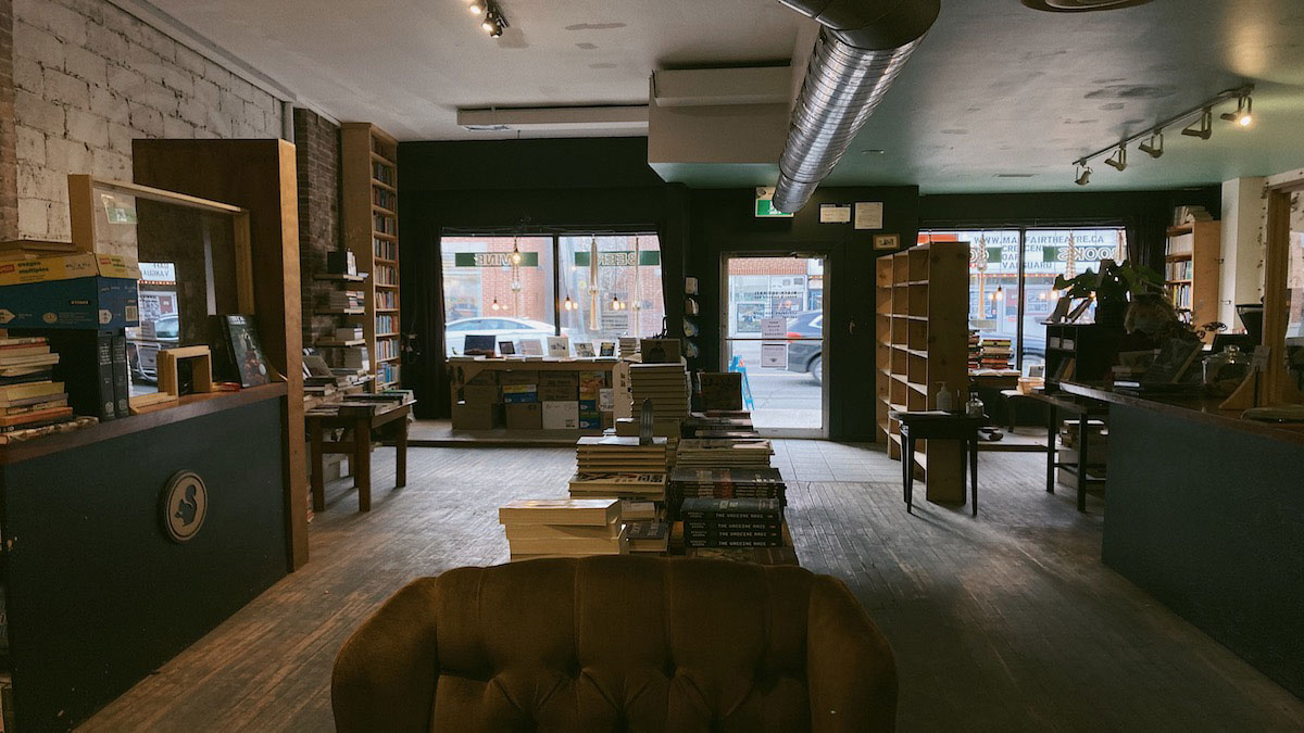 Front space of Black Squirrel Books, with counters on right and left, a long table with books down the middle with aisles on either side, and front store windows at the back of the photograph.