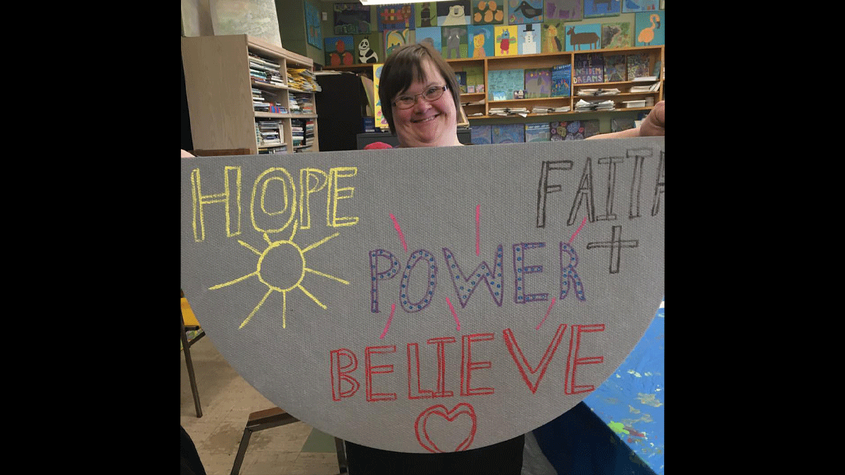 Photo of Analisa Kiskis holding a banner that says "HOPE POWER FAITH+BELIEVE ME"