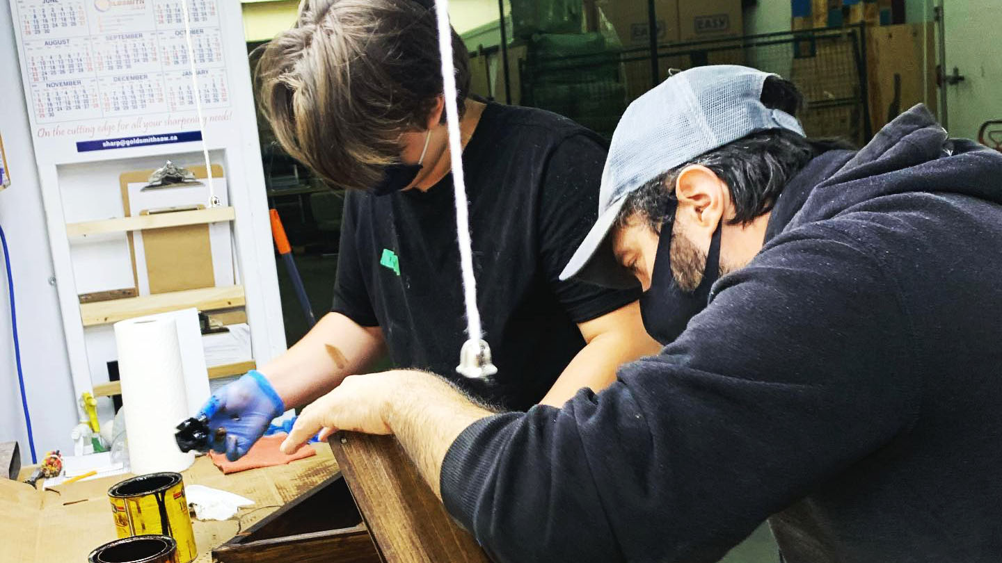 Two men are wearing masks and leaning over a project, staining the wood.