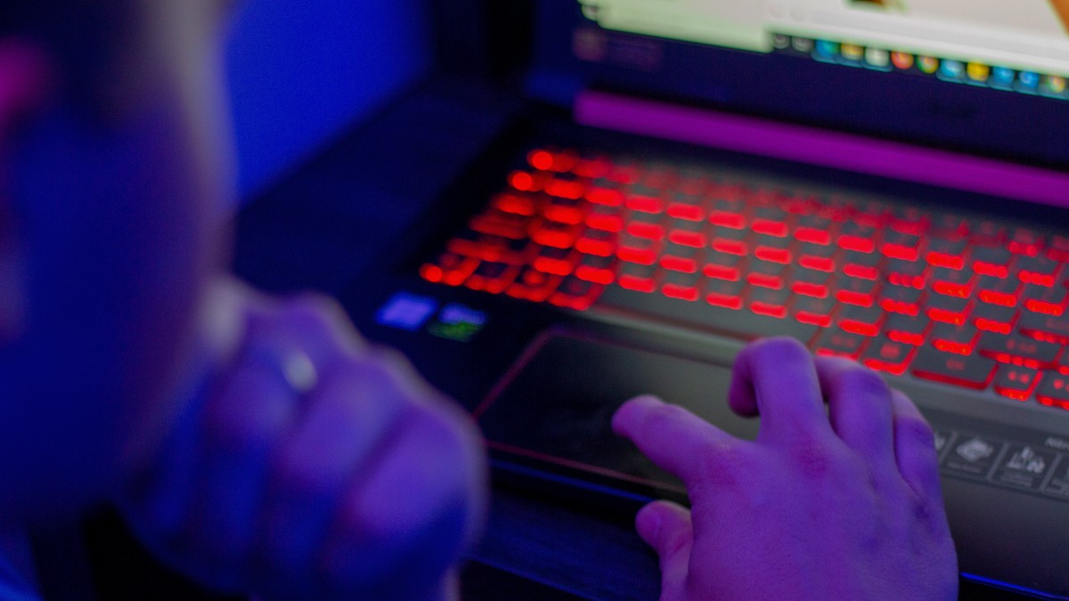 A person looking at their laptops screen and using their hand to move the mouse. The room is dark and the keyboard is backlit with red light.