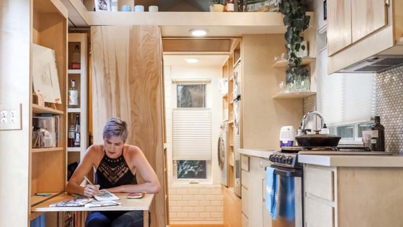 In this photo, Kari Gale, owner of a tiny home, sits and writes at her small fold out desk in her tiny kitchen with light wood cabinets and modern appliances.