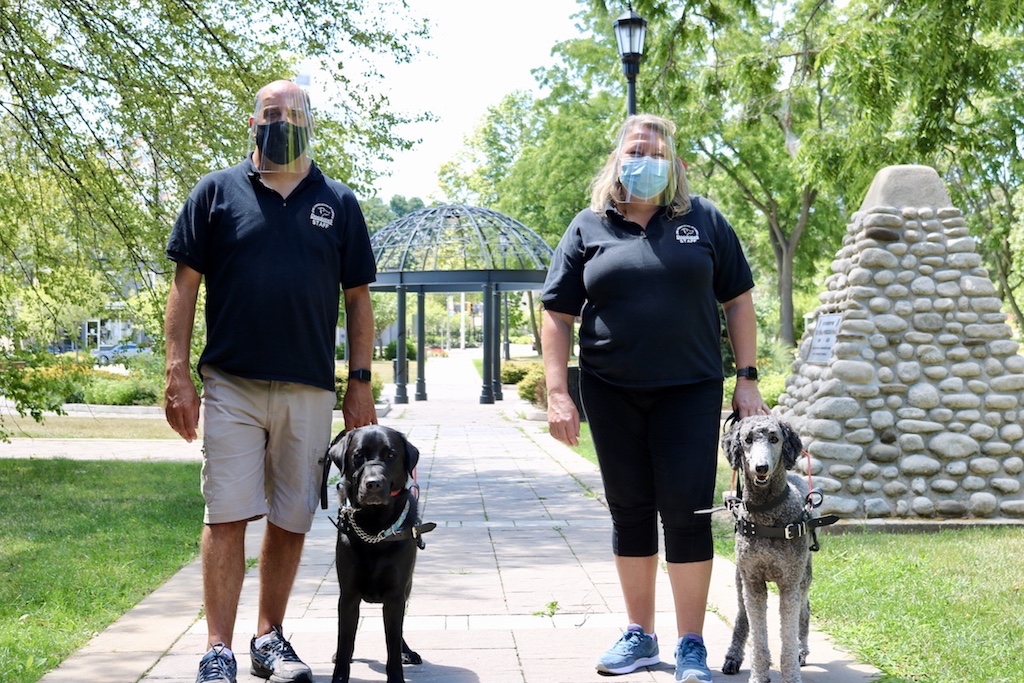 Two Lions Foundation dog trainers, wearing surgical face masks and plastic face shields, pose in an outdoor park setting with two guide dogs standing leashed at their sides.