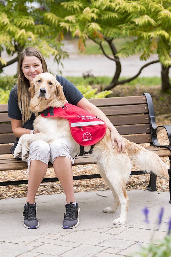 A woman sits smiling on a park bench outdoors with an autism assistance dog guide standing with its front paws on her lap. 