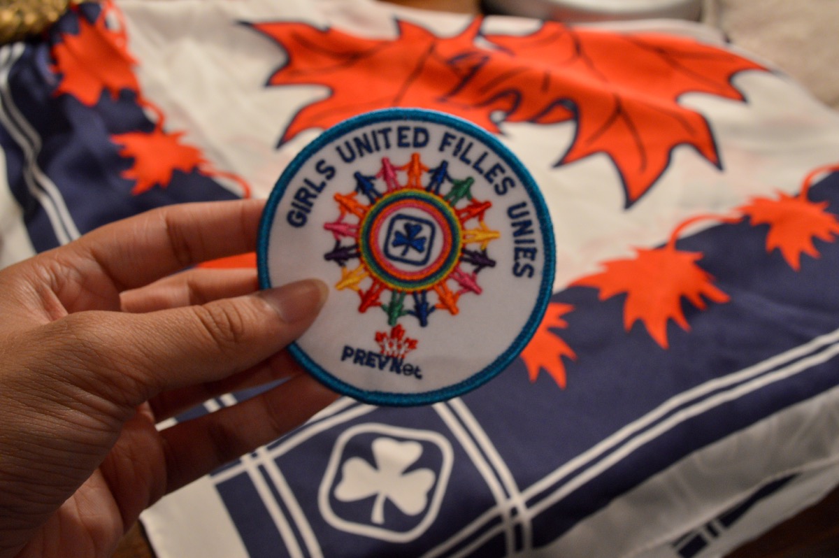 Hand holding a circle badge that says "Girls United Filles Unies" with a screw in the background with the Girl Guide emblem and red maple leafs