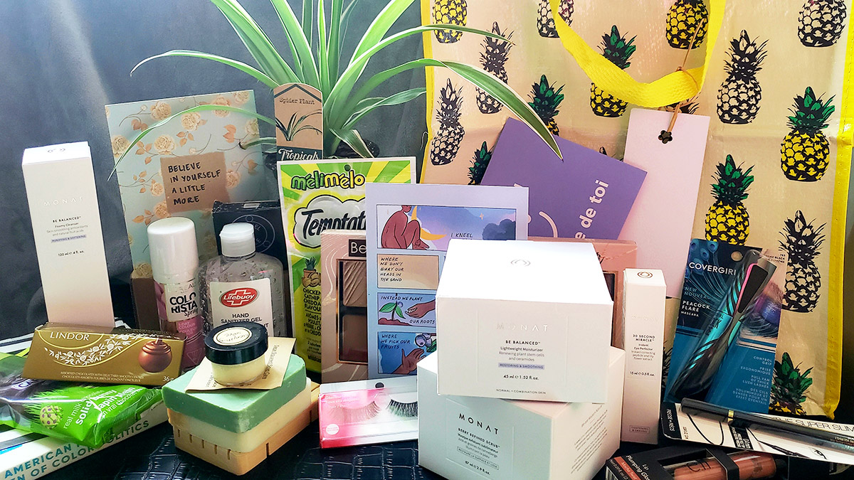 Colourful care package contents laid out on a table. Items include makeup products, a plant, hair products, chocolate, a customized card and a large, yellow reusable bag with a pineapple pattern.