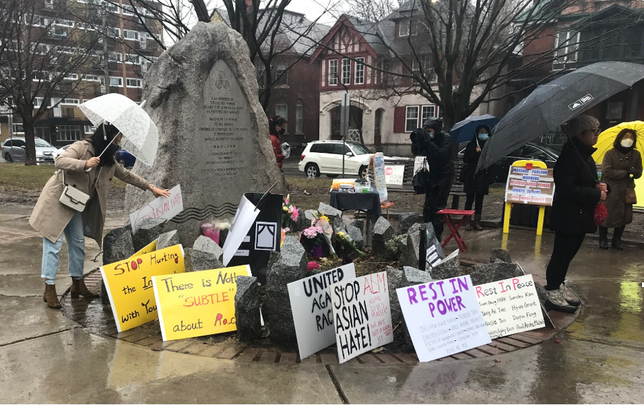 Scene shows stone monument surrounded by hand-made signs with woman under umbrella leaning forward to add her sign