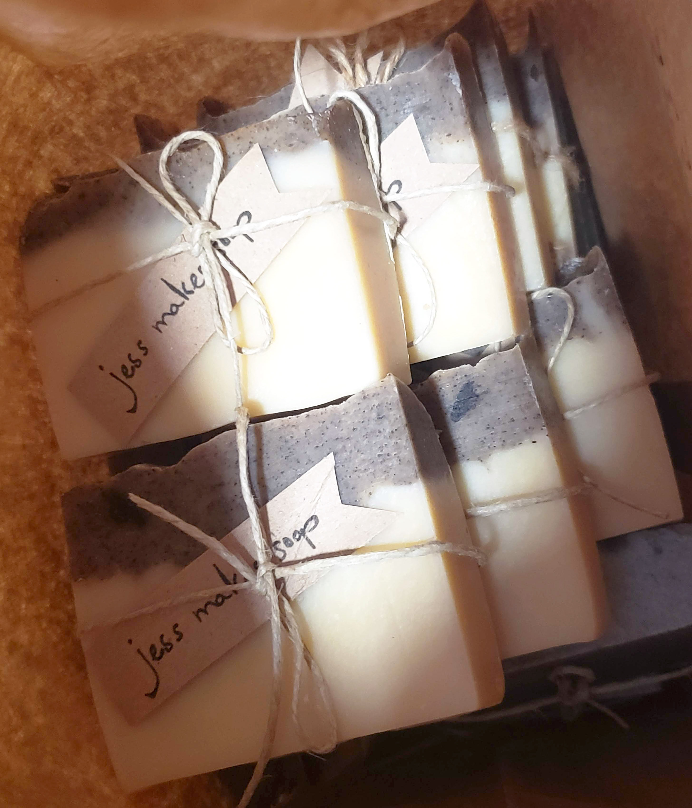 Stacked bars of homemade vanilla soap in a paper bag. The bars are ivory coloured, with a grey and black-speckled top layer. Each bar is tied with a piece of twine, holding a brown label with the black handwritten words "jess makes soap."