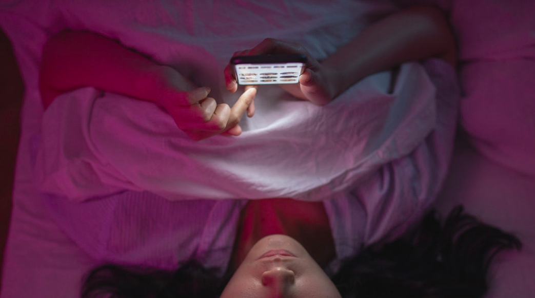 A young woman is show lying in bed looking at her smartphone