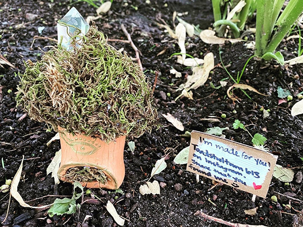 A clay toadstool covered in moss and painted beige sits in a garden, next to a small sign that says, "I'm made for you! Toadstooltown613 on Instagram. Share that you found me."