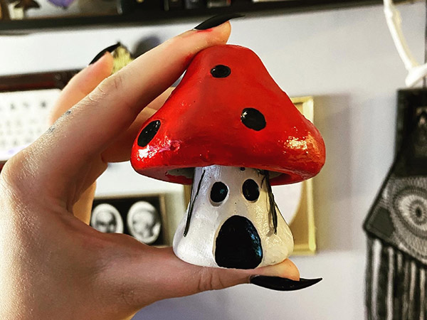 A hand with black fingernail polish holds up a small toadstool sculpture, painted white, red and black.