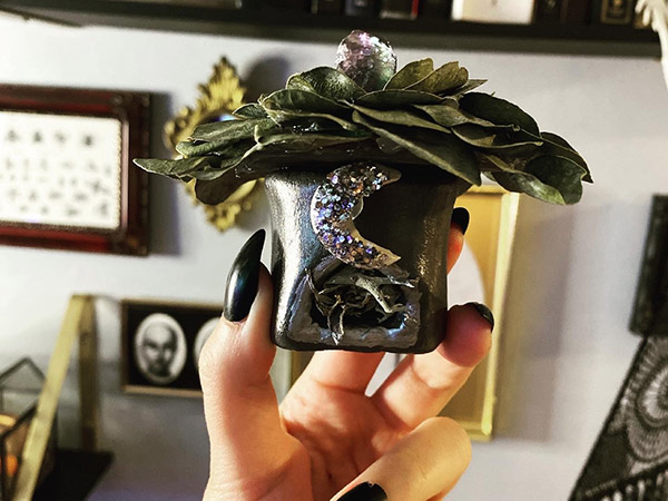 A hand with black fingernail polish holds a small clay toadstool sculpture, painted black and decorated with purple crystals and dry leaves.