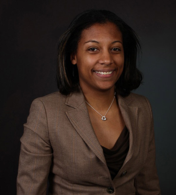 A woman wearing a brown suit smiles and stands in front of a grey background.