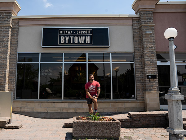 A man in a red t-shirt stands in front of a gym building with a sign that reads "Ottawa Crossfit Bytown".