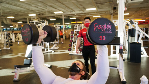 A man lifts two 50 pound dumbbells while a personal training watches from behind.