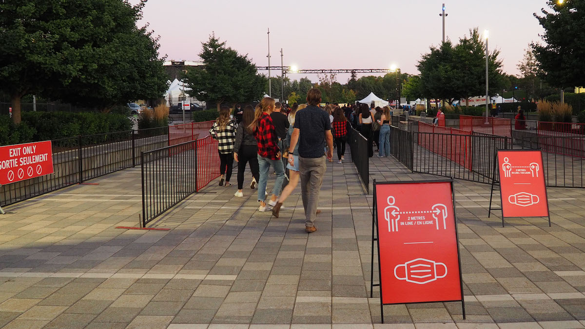 Attendees walk towards the entrance of an outdoor concert event, red signs displaying social distancing rules and mask mandates are in the forefront. 