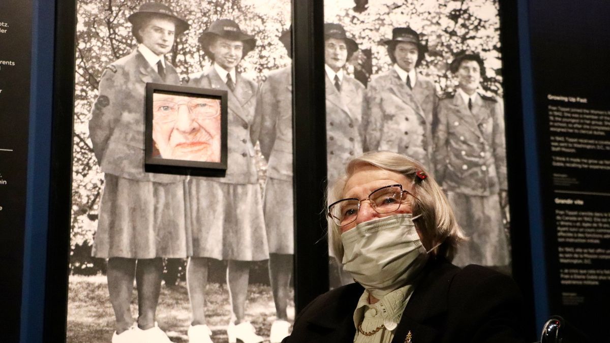 An older woman in a wheelchair and mask sits in front of a painted portrait of her. Behind the portrait is a large-scale photo of a group of women in uniform during the Second World War.