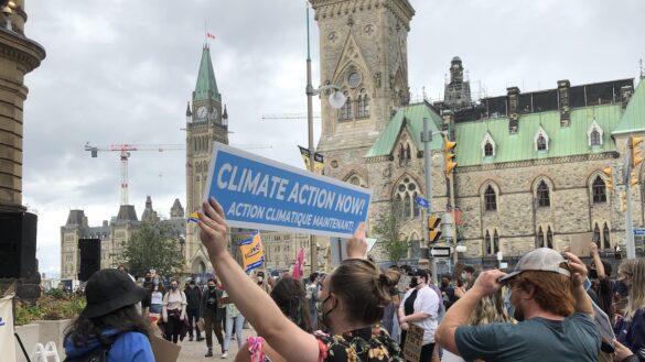Protestor holding a sign that says "Climate action now. Action Climatique maintenant" with the Parliament in the background.