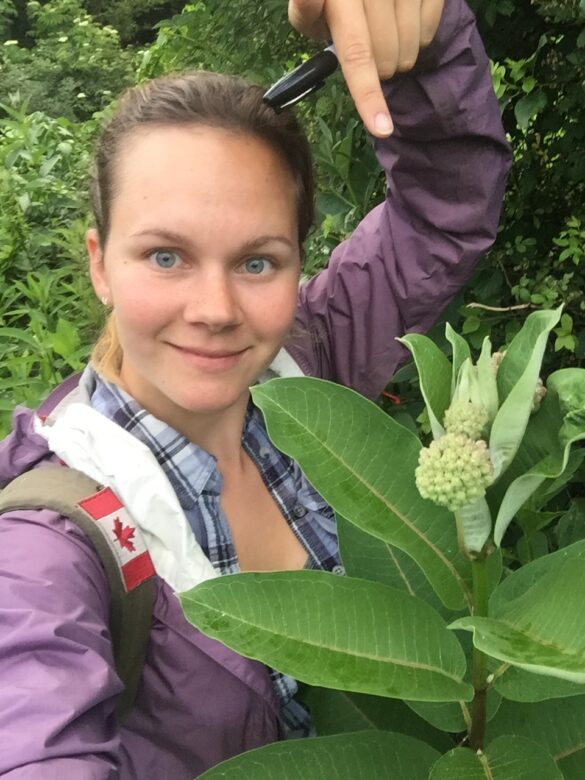 Megan Reich stands outdoors pointing to the flower of a milkweed plant she is holding. The photo was taken during her trip across the eastern United States.