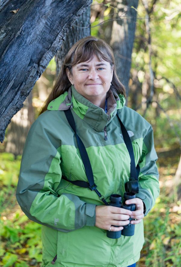 Nicola Koper, a middle-aged woman stands in a forest in a green jacket. She is holding a pair of binoculars and smiling.