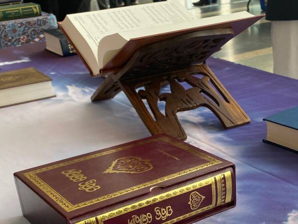 A red Qur'an with gold lettering in the foreground, and an open Qur'an in the background, on a table display.
