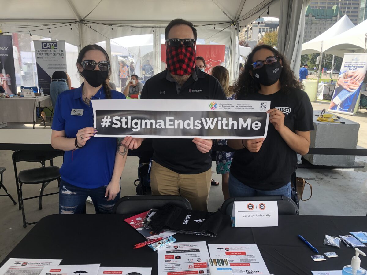 Image shows three people holding "stigma ends with me" banner at the Carleton University table outside of Ottawa City Hall.