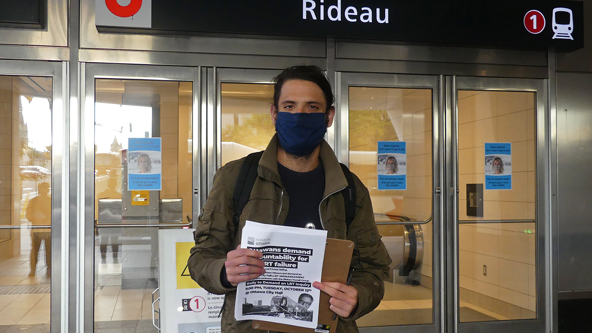 Sam Hersh, a board member with Horizon Ottawa, stands in front of the closed doors at Rideau station. He is wearing a blue mask and a green jacket while holding up flyers for an upcoming rally event.