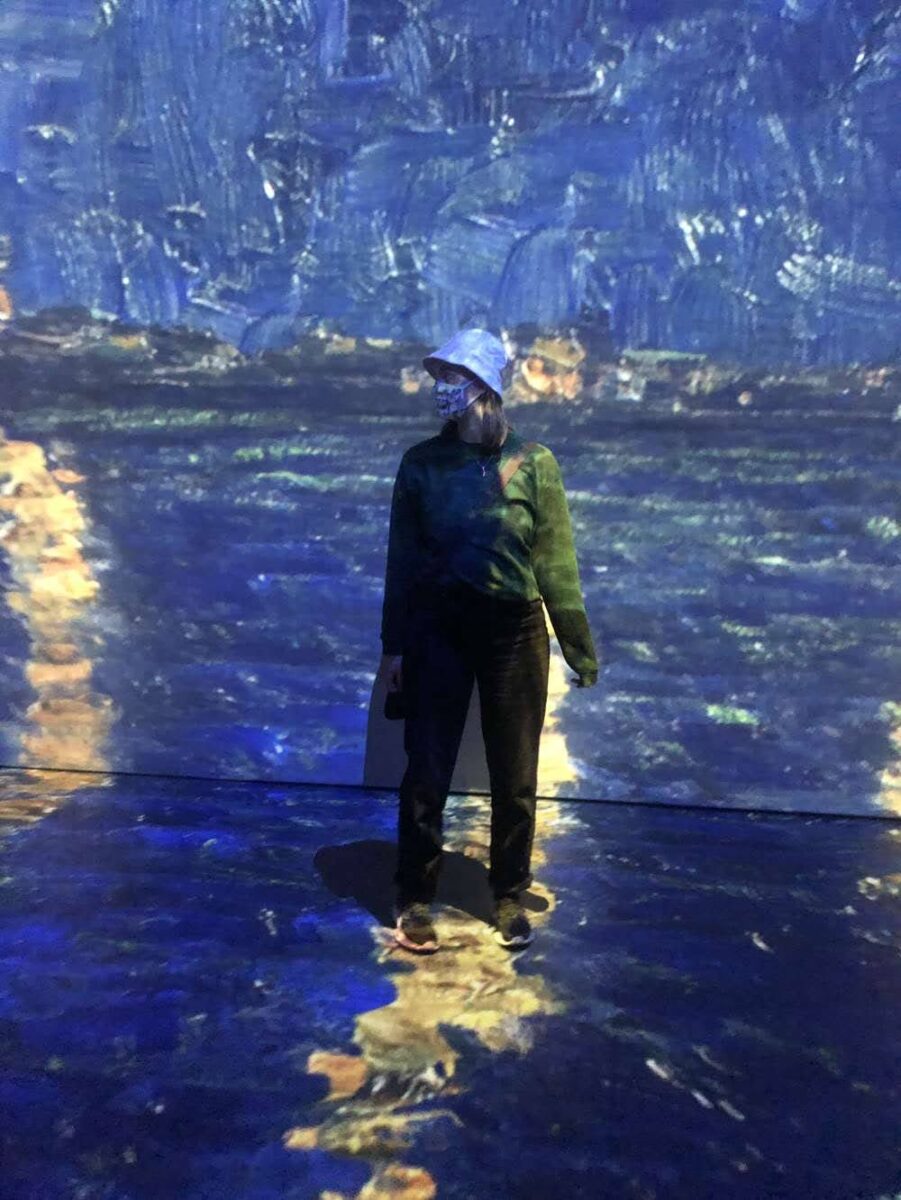 Person standing in a projection of a blue and yellow painting at Beyond Van Gogh: The Immersive Experience
