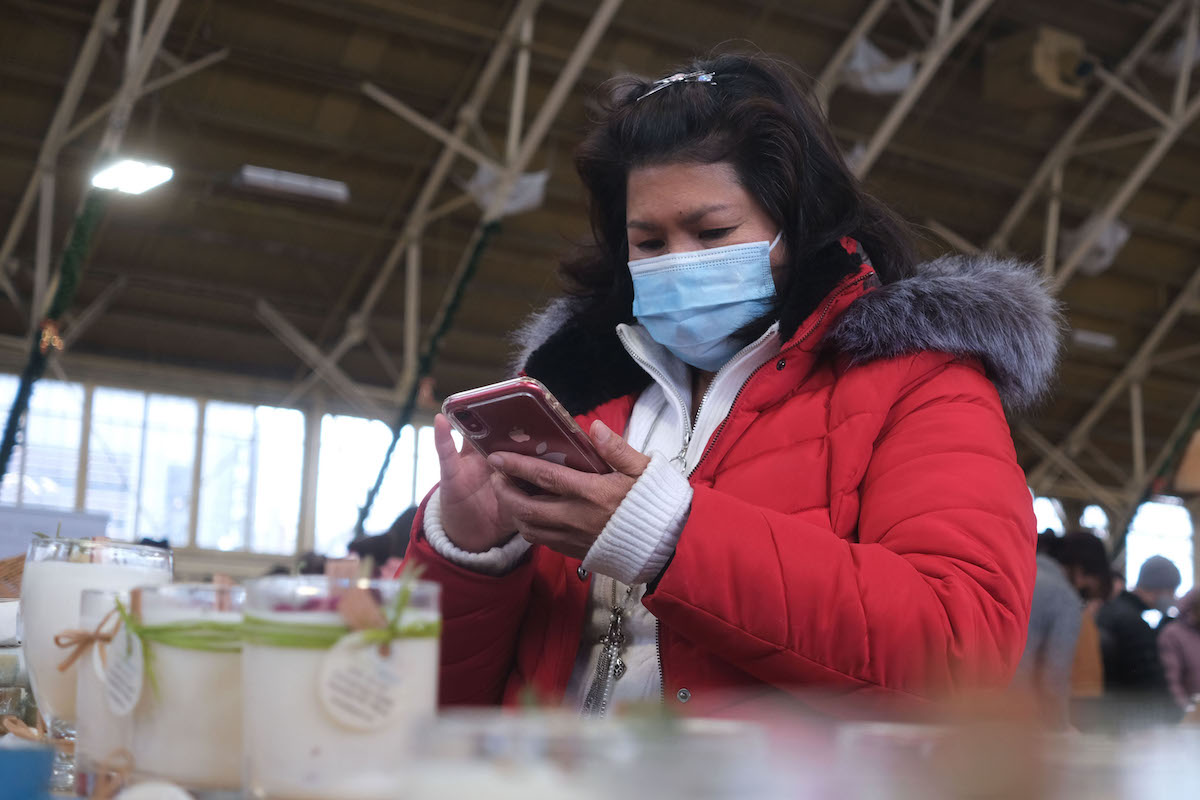 Woman in red coat stands behind a table full of candles and looks down at her phone at the Christmas market.