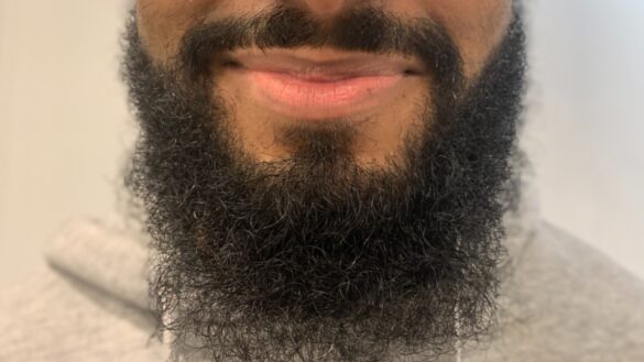 Half of a face is shown. Person has a darker skin tone and dark moustache that connects to large beard.