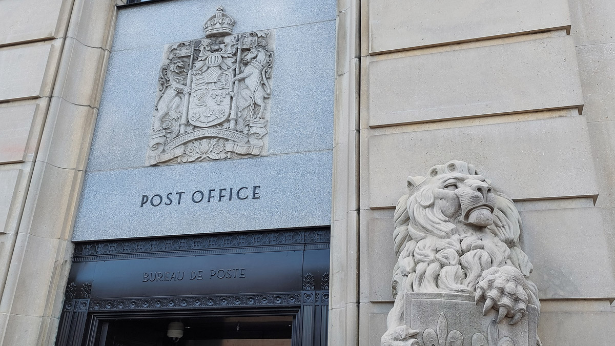 A Canadian coat of arms in grey stone sits above a doorway on the wall. To the right, a statue of a lion holding a shield with three fleurs-de-lis. 