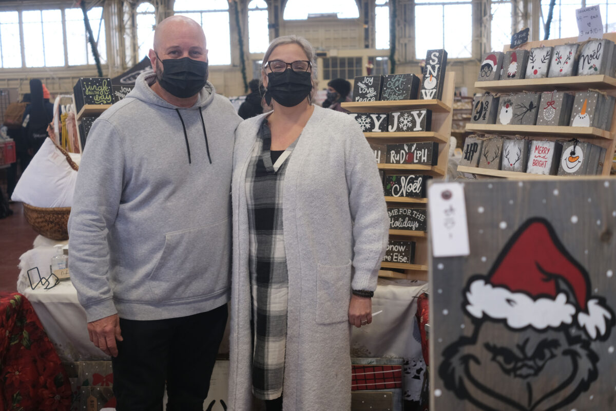 Woman and man are dressed in grey and embracing each other standing in front of dozens of hand painted Christmas signs at the Christmas market.