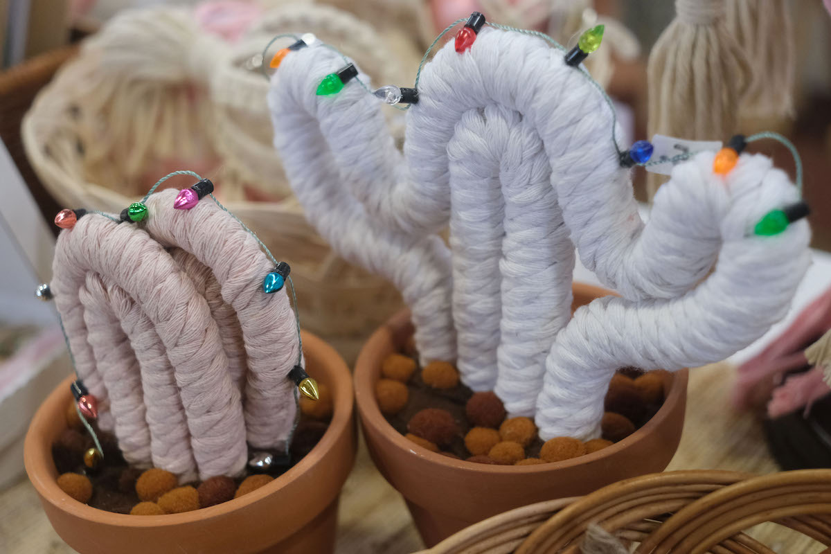 Two cacti made out of macrame are in brown pots. One is pink, the other is white, and both are decorated with small Christmas lights.