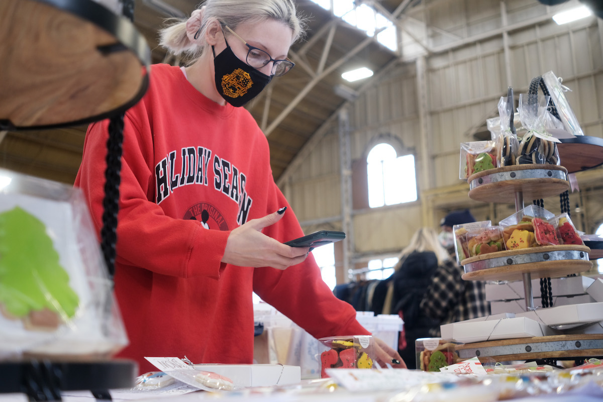 Woman in a red sweater that reads holiday season is standing at a table at the Christmas market surrounded by cookies. She is looking down at her phone.