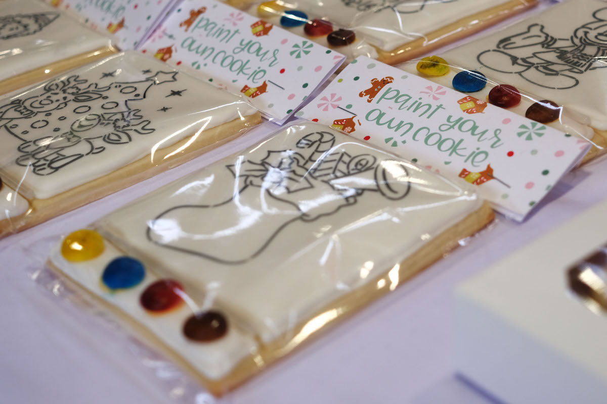 A table with an assortment of cookies. The white cookies are square shaped and have an outline of a stocking on them. Underneath the cookie are four colours: yellow, blue, red, and purple. for customers to paint their own cookies. On each cookie is a label that says paint your own cookie/