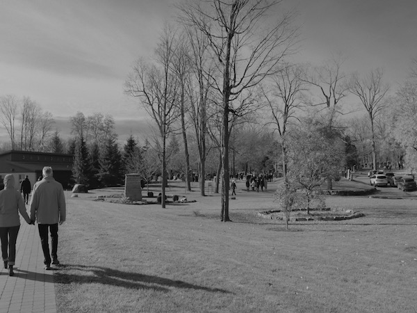 In black and white, a couple holds hands walking out of the ceremony, in the distance more people are leaving the grounds. 
