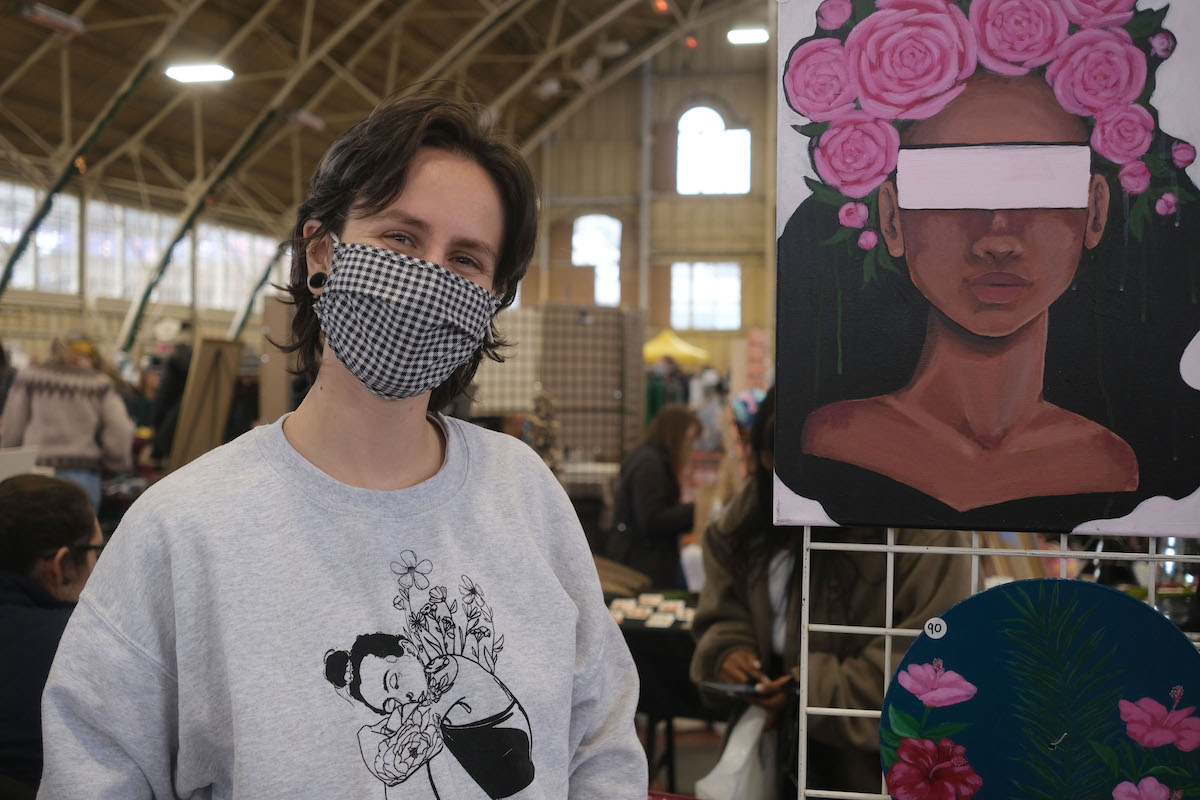 Vendor with short brown hair is smiling beside two paintings at the Christmas market. The top painting has a woman with flowers in her hair and her eyes are covered. The bottom painting is a blue background with pink flowers.