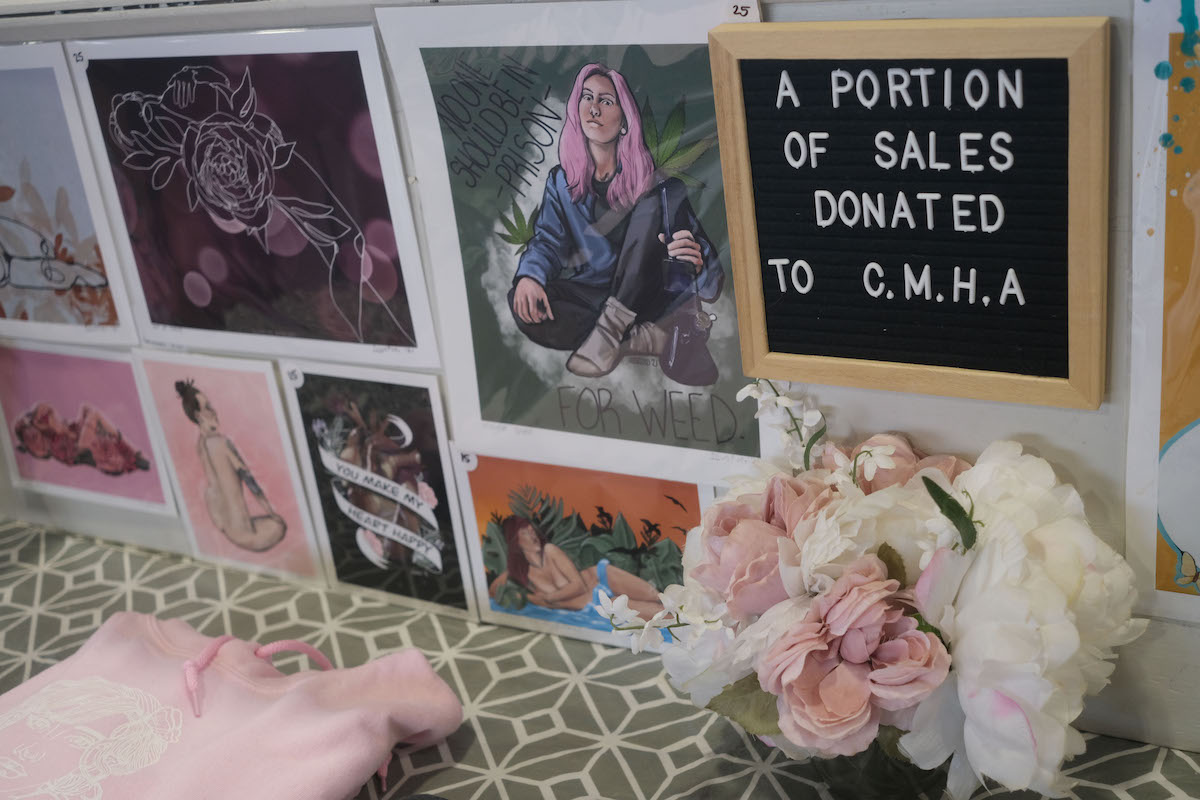 An assortment of prints are lined up. There is a pink sweater folded beside pink and white flowers. In focus is a sign that says a portion of sales donated to CMHA.