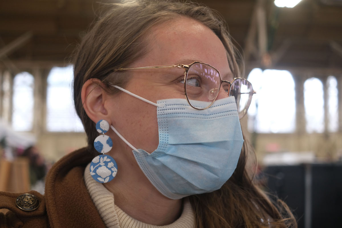 Woman with long brown hair and glasses looks to the side. She is wearing long blue and white earrings.