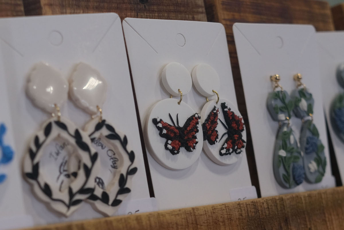 Three pairs of handmade earrings are lined up at the Christmas market. The earrings in focus are white, and have orange butterflies on them.