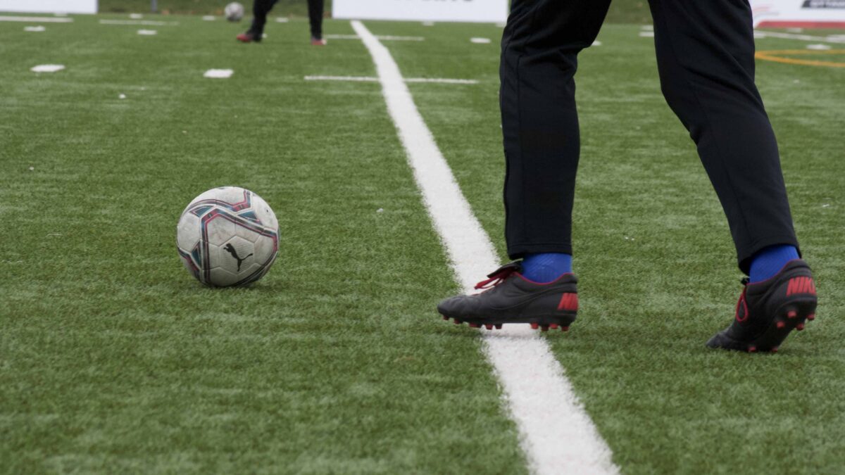 A soccer player runs up to pass a ball to their teammate on an artificial turf field.