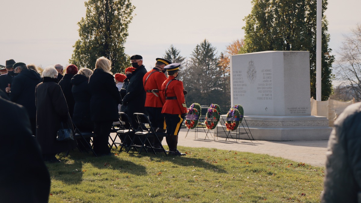 At the front of the VIP section stand two military personnel in red suits, bright against the pale background. Wreaths have been set in front of a stone monument.